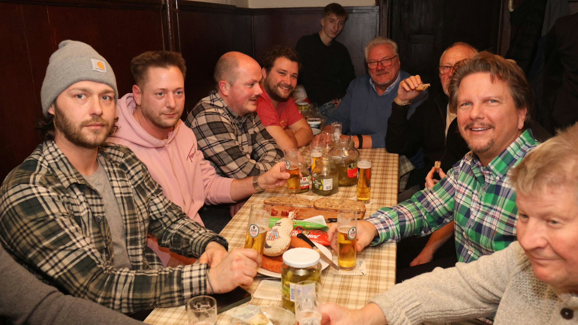 Eine Männerrunde sitzt an einem langen Tisch und spielt Dobbeln. Auf dem Tisch stehen Biergläser und Teller, auf denen Wüste liegen.