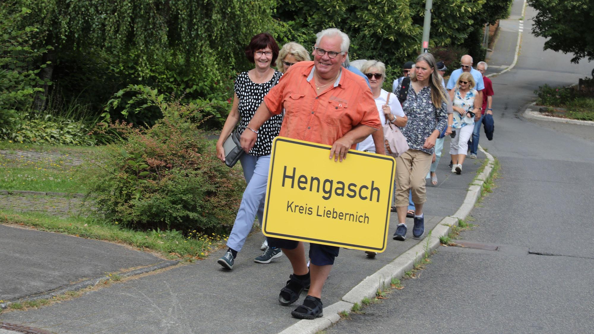 Michael Iwanzik mit Hengasch-Ortsschild und einigen Menschen im Gefolge.