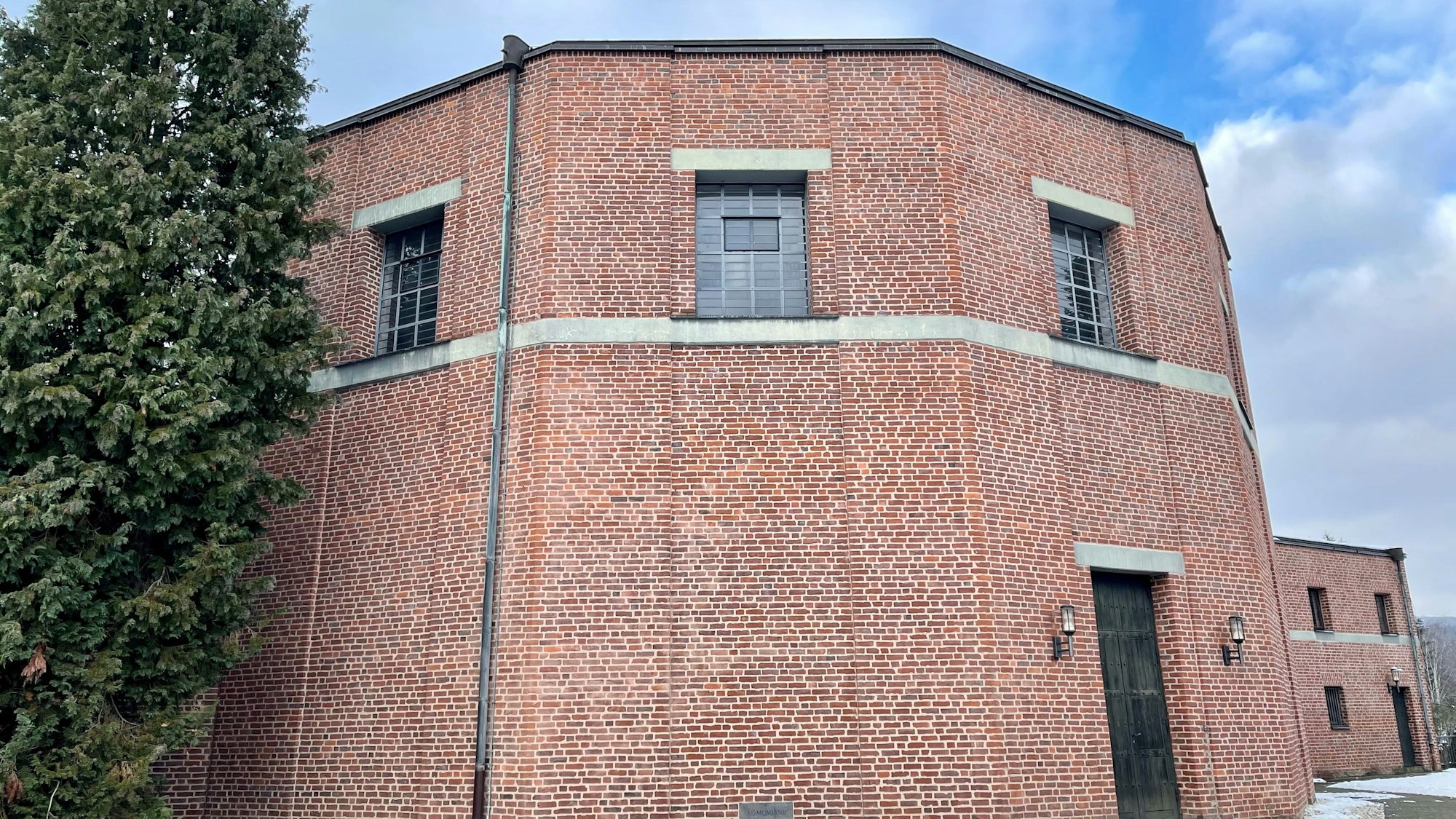 Ein nahezu runder Fachwerkbau mit dunkler Tür und Fenstern nur im oberen Bereich steht in winterlicher Landschaft.