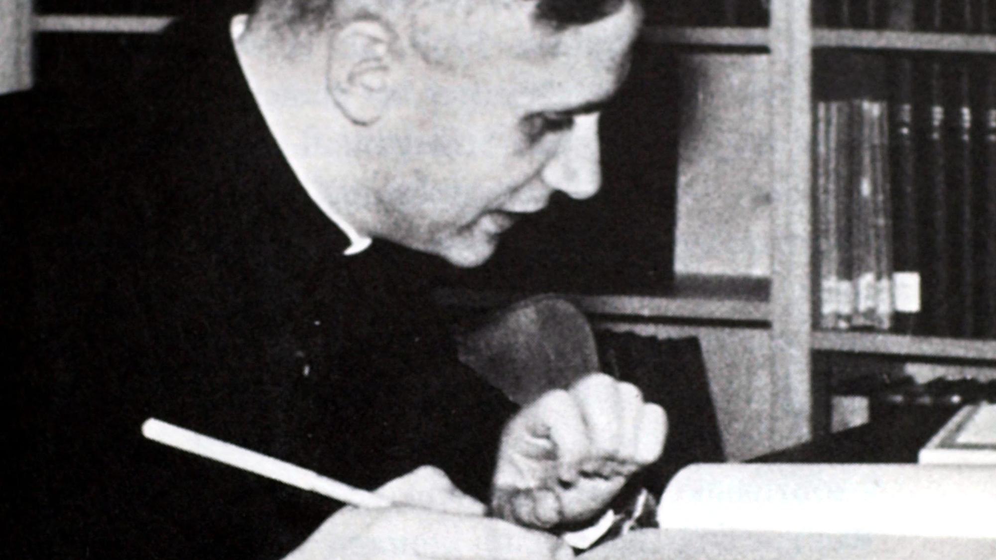 Ein altes Schwarz-Weiß-Foto zeigt einen jungen Mann, der mit einem Stift in der Hand an einem Tisch mit aufgeschlagenen Büchern sitzt.