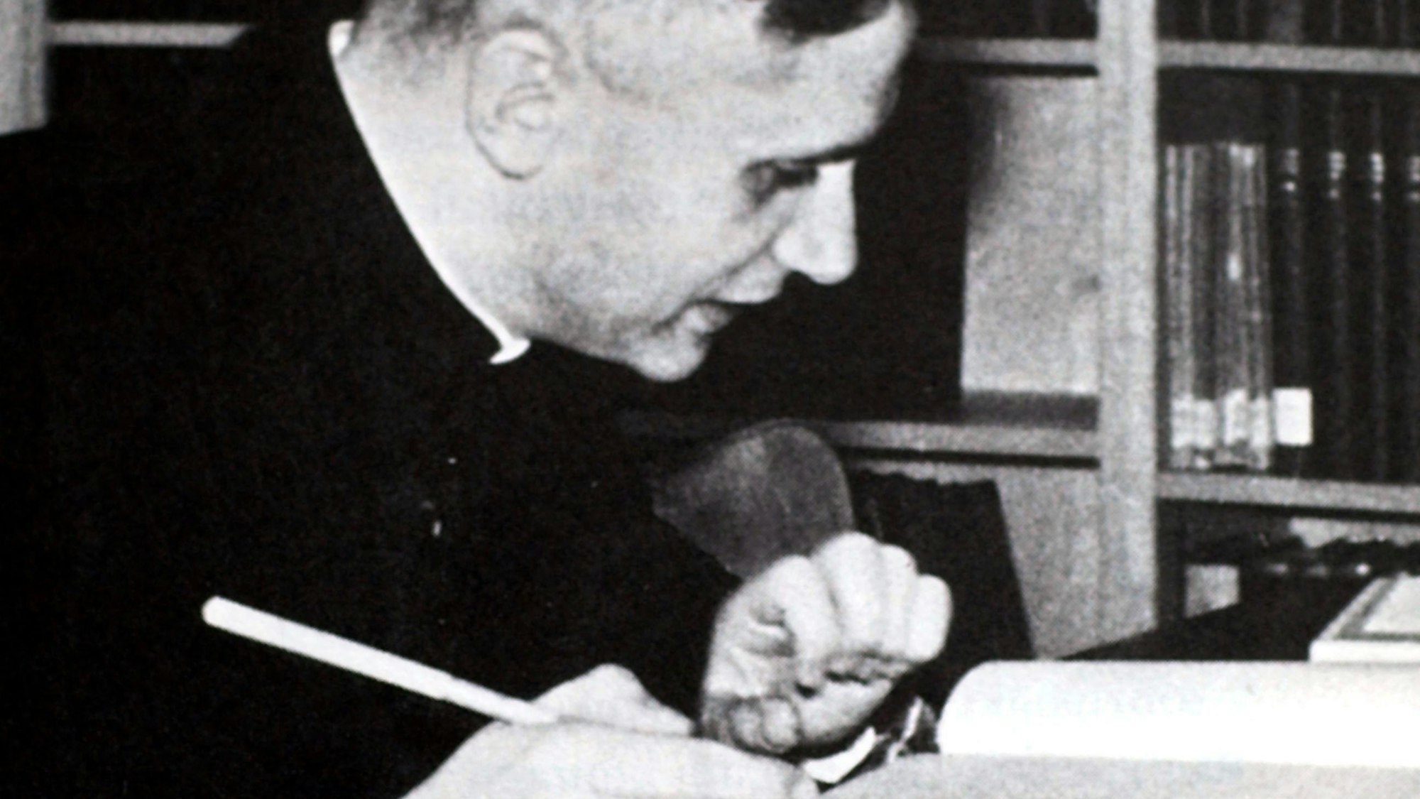 Ein altes Schwarz-Weiß-Foto zeigt einen jungen Mann, der mit einem Stift in der Hand an einem Tisch mit aufgeschlagenen Büchern sitzt.