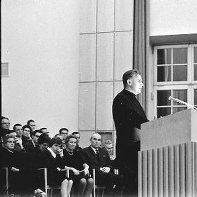 Ein altes Schwarz-Weiß-Foto zeigt einen schwarz gekleideten Mann an einem Rednerpult mit Mikrofon, hinter ihm sitzen mehrere Menschen.