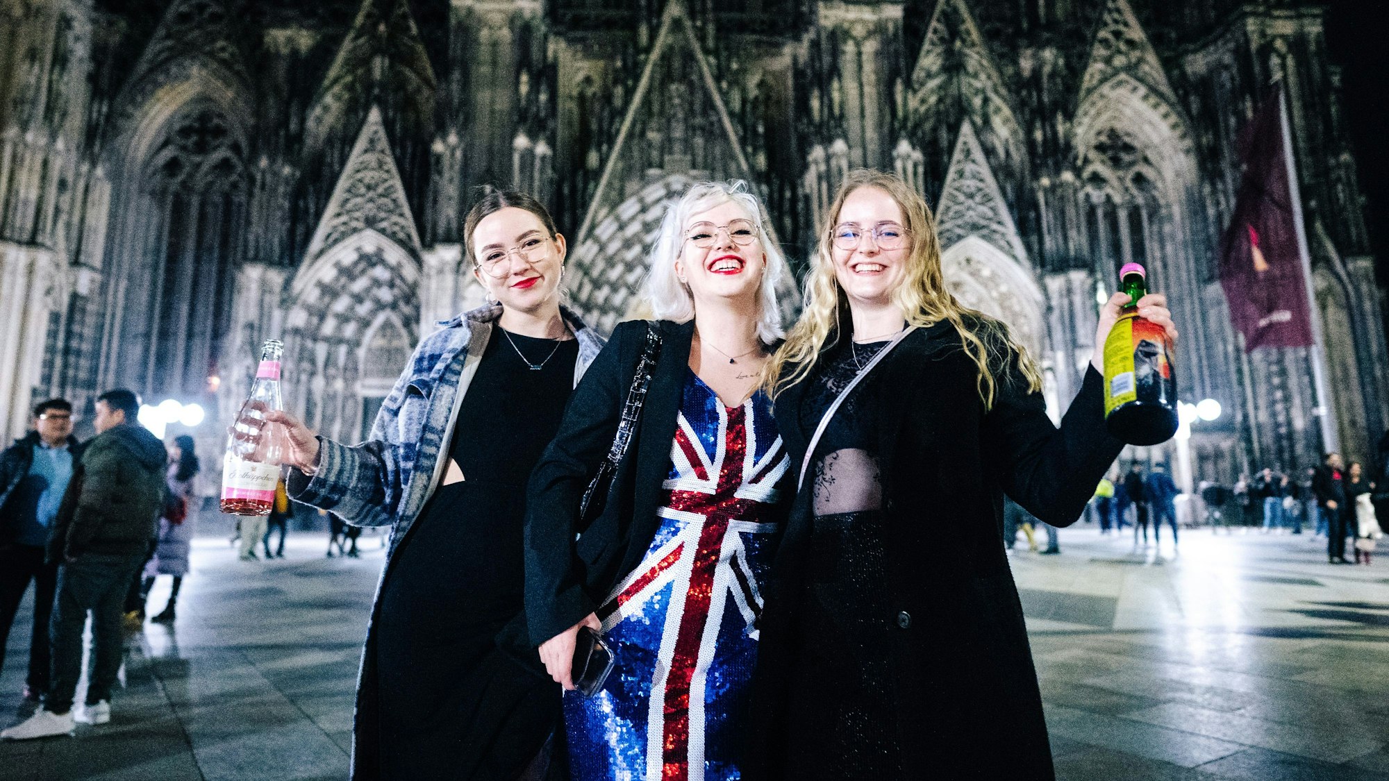 Drei junge Damen in Feier-Outfits lächeln vor dem Dom in die Kamera und halten ihre mitgebrachten Sektflaschen hoch.