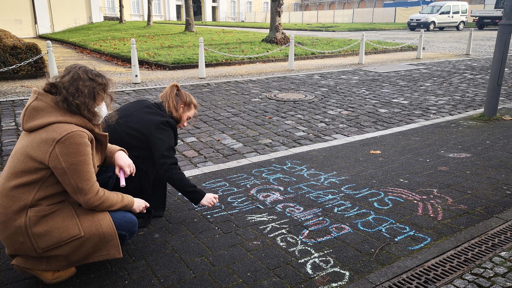 Zwei junge Frauen schreiben mit bunter Kreide einen Aufruf auf den Fußweg, mit dem sie dazu auffordern, Erfahrungen von Belästigungen in der Öffentlichkeit zu teilen.