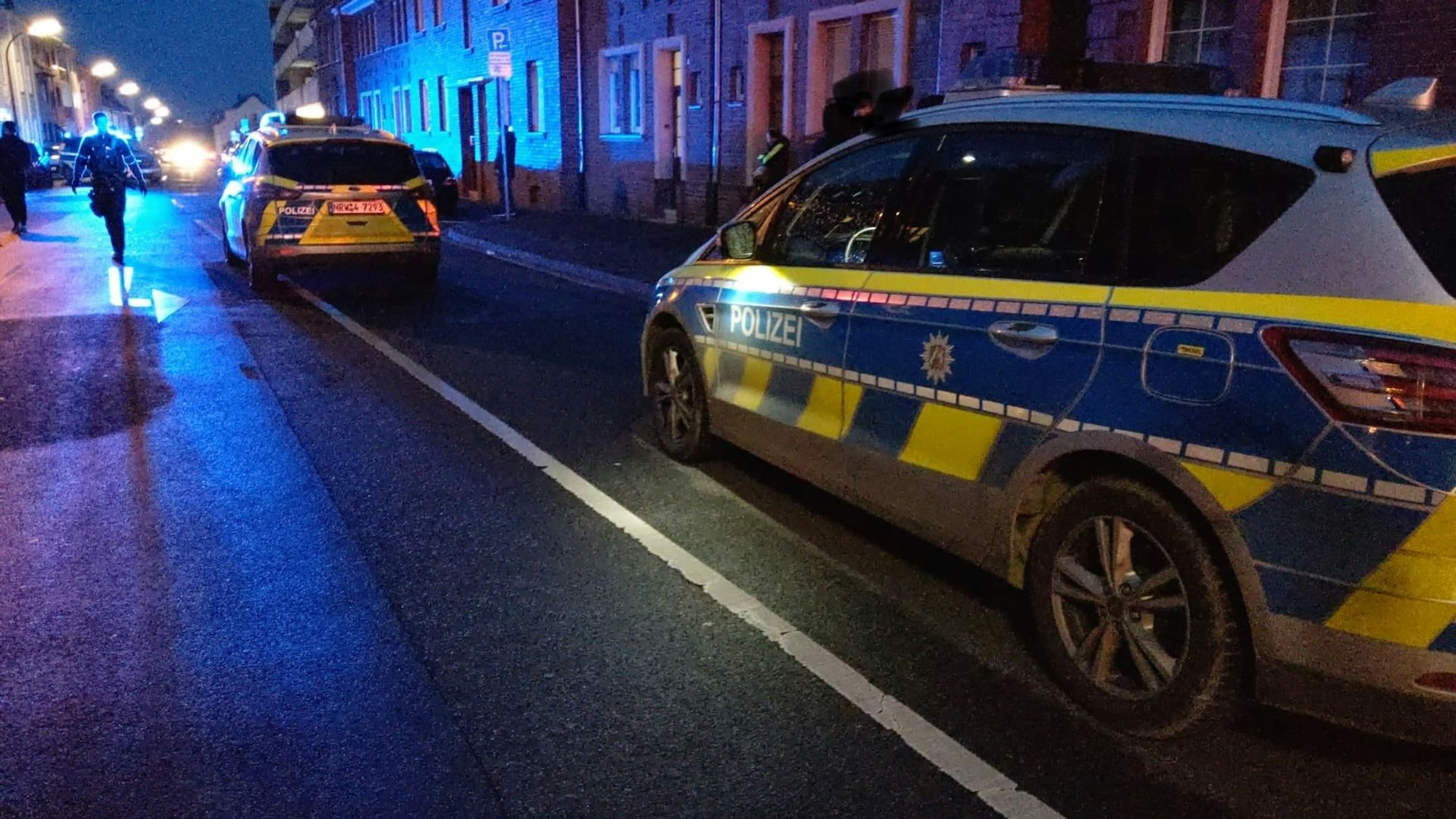 Zwei Streifenwagen stehen hintereinander auf einer Straße, man sieht mehrere Polizisten im Hintergrund. Die Straße ist dunkel, bis auf Laternen und Blaulicht.