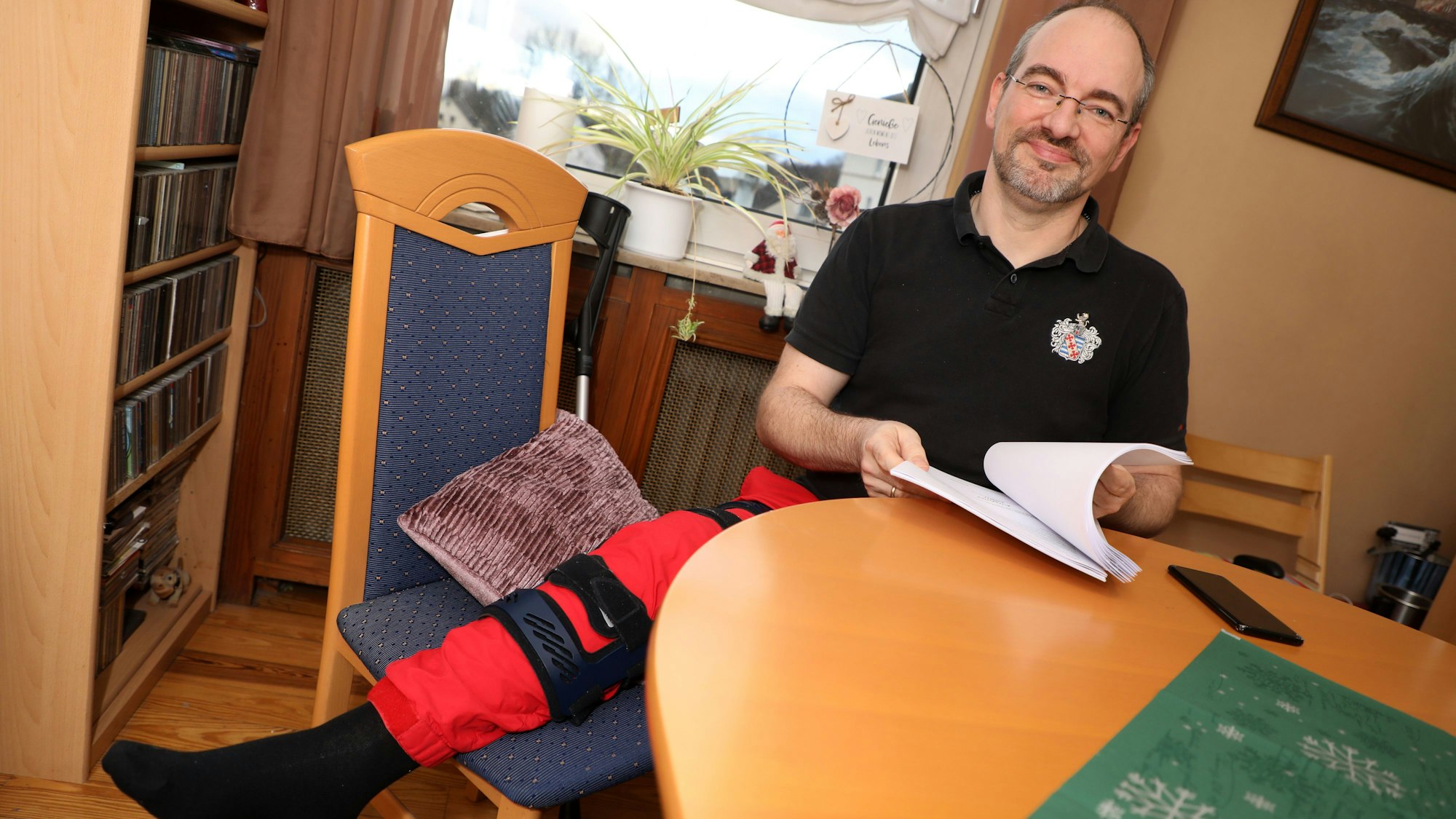 Alexander Pfister sitzt an einem Tisch und blättert in Dokumenten, sein rechtes Bein ist geschient und liegt hoch.