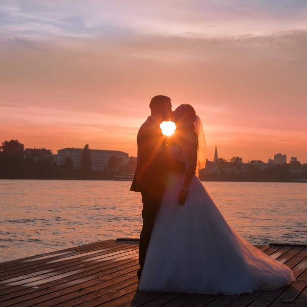 Ein Hochzeitspaar steht im Sonnenuntergang auf einem Steg vor einem Fluss.