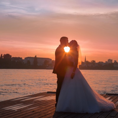 Ein Hochzeitspaar steht im Sonnenuntergang auf einem Steg vor einem Fluss.