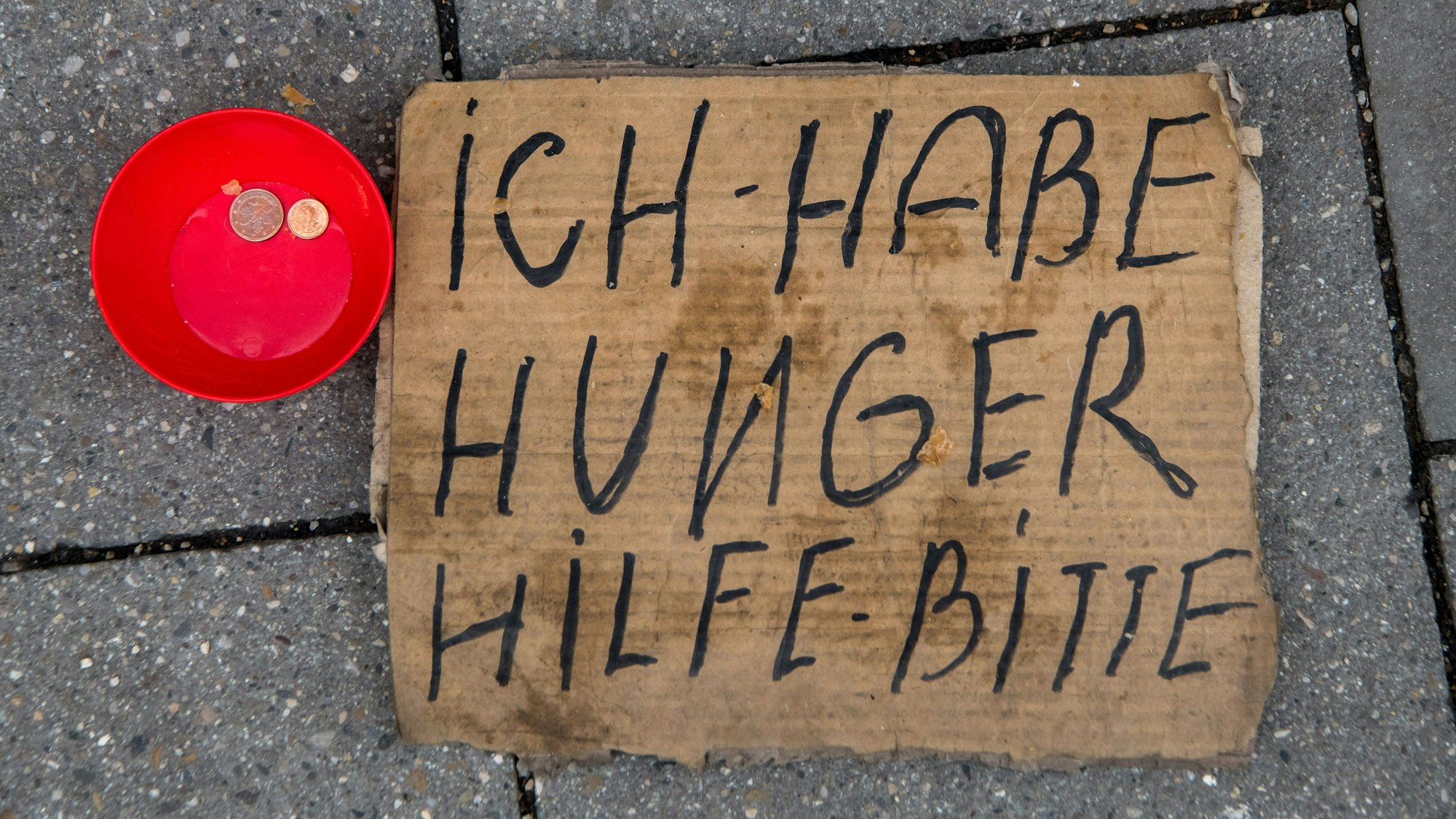Ein dreckiges Pappschild mit der Aufschrift Ich habe hunger Hilf - bitte, liegt auf der Straße. Daneben steht eine rote kleine Schüssel mit zwei Münzen drinnen.