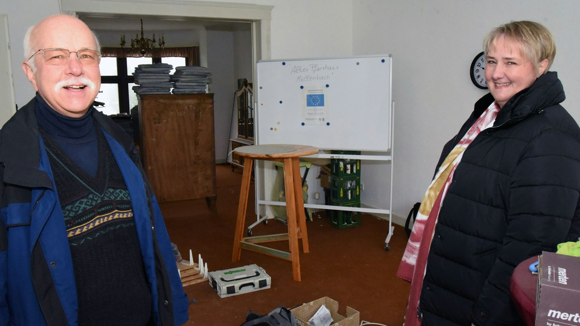 Anke Bockelmann und Georg-Wilhelm Overbeck stehen in einem der renovierungsbedürftigen Räume des Pfarrhauses in Marienheide-Müllenbach. Im Hintergrund sind weggerückte Schränke und gepackte Kartons zu sehen.