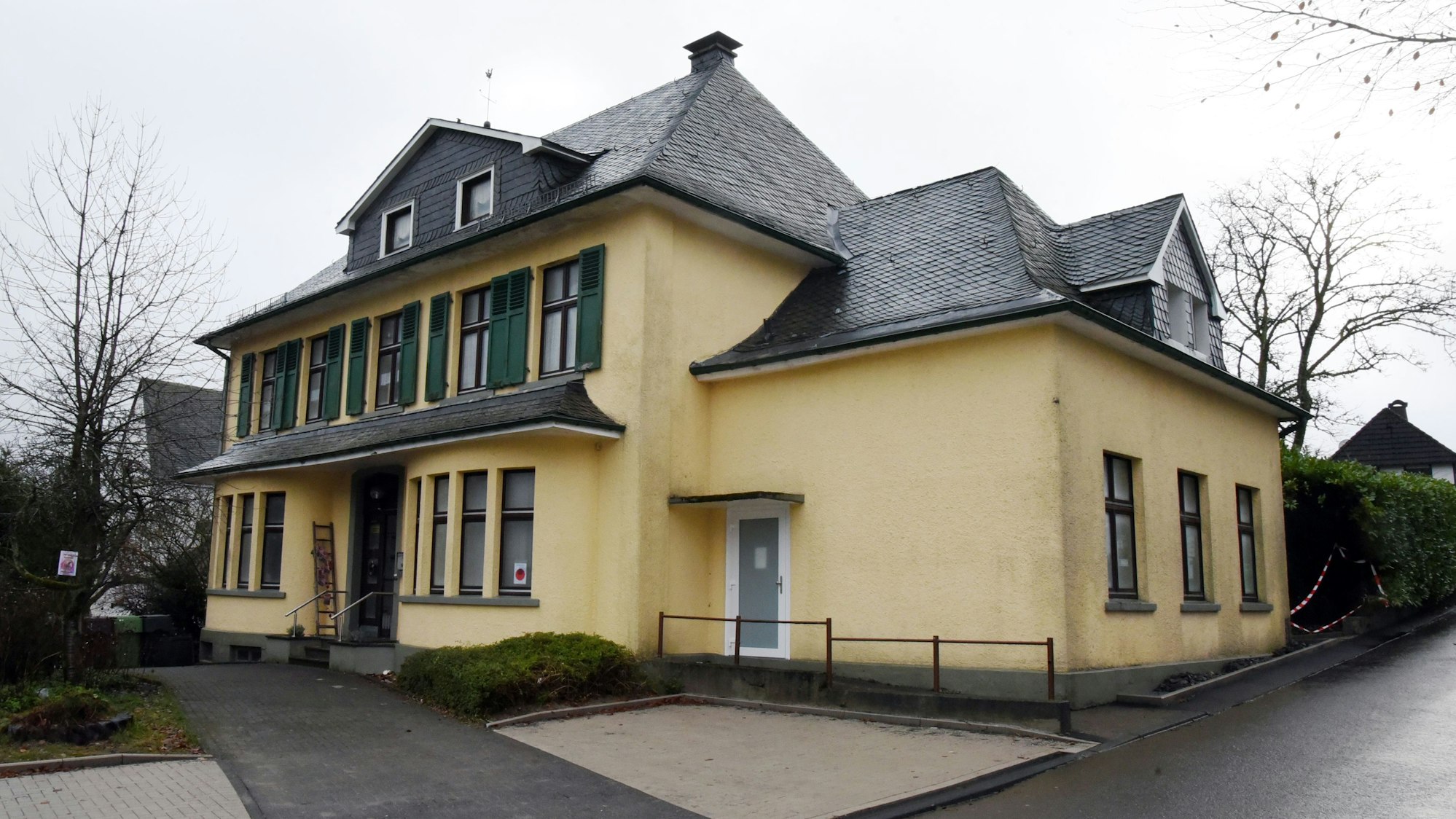 Das Pfarrhaus in Marienheide-Müllenbach ist als Außenansicht zu sehen. Das Gebäude ist gelb gestrichen, mit grünen Fensterläden in der oberen Etage.