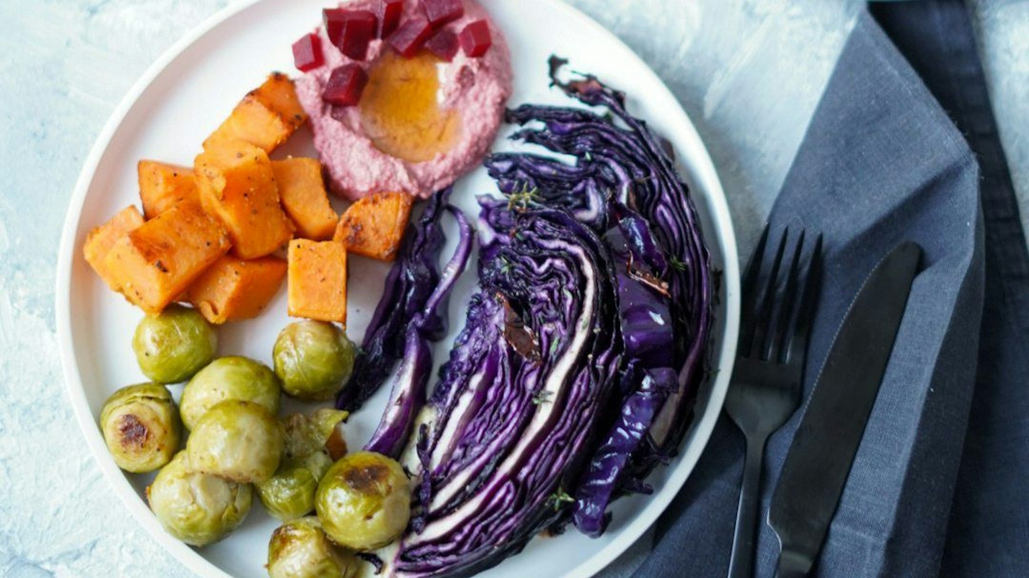 Wintergemüse, bestehend aus Rotkohl, Rosenkohl, Süßkartoffel und rötlichem Hummus, farbenfroh auf einem Teller angerichtet.