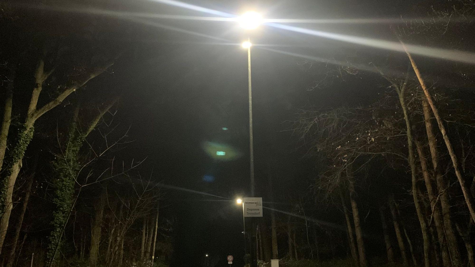 Eine Laterne strahlt in der Dunkelheit heller als der Mond dahinter. Links und rechts stehen Bäume.