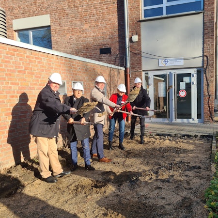 Fünf Männer mit Bauhelmen stehen vor dem Krankenhaus auf Baustellenboden und haben einen Spaten i der Hand.