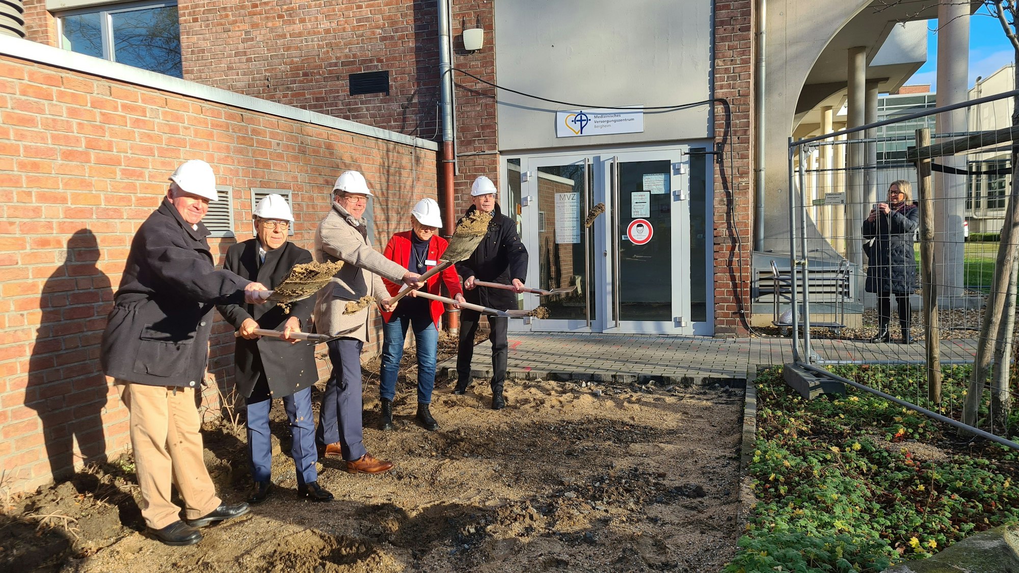 Fünf Männer mit Bauhelmen stehen vor dem Krankenhaus auf Baustellenboden und haben einen Spaten i der Hand.