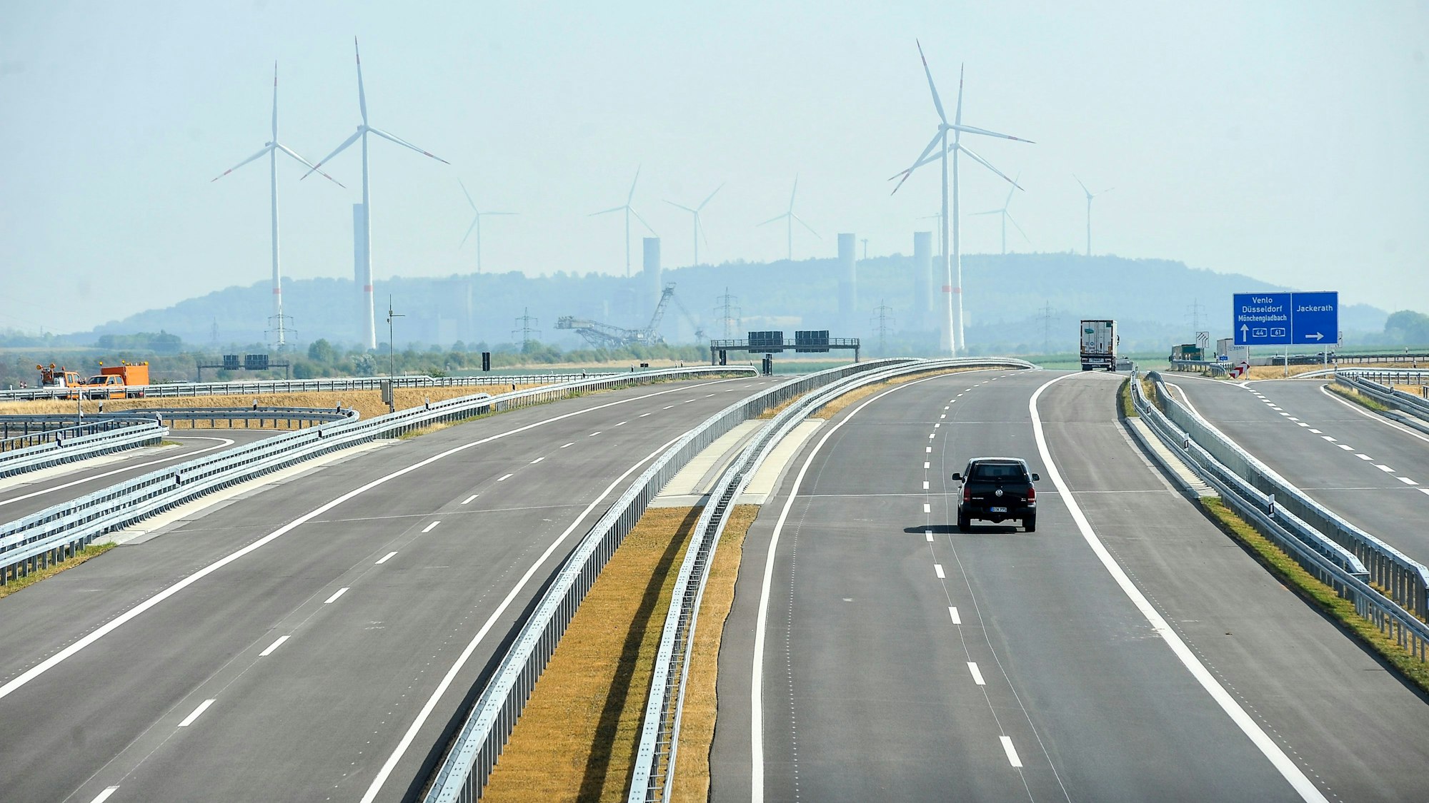 Ein Auto fährt auf einer Autobahn. Im Hintergrund sind Windräder und der obere Teil eines Braunkohle-Baggers zu sehen.
