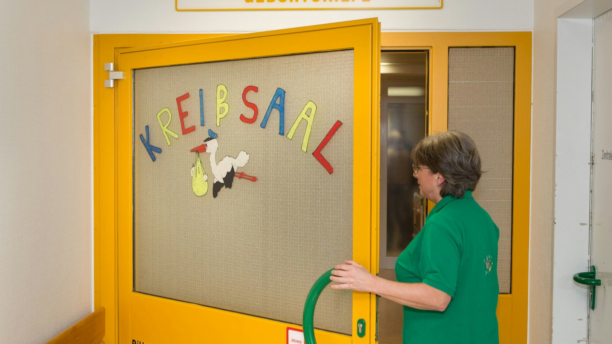 Eine Hebamme betritt auf der Geburtshilfestation im Krankenhaus Gummersbach einen Kreißsaal. Auf dem Zugang klebt ein aus Papier ausgeschnittener Storch.