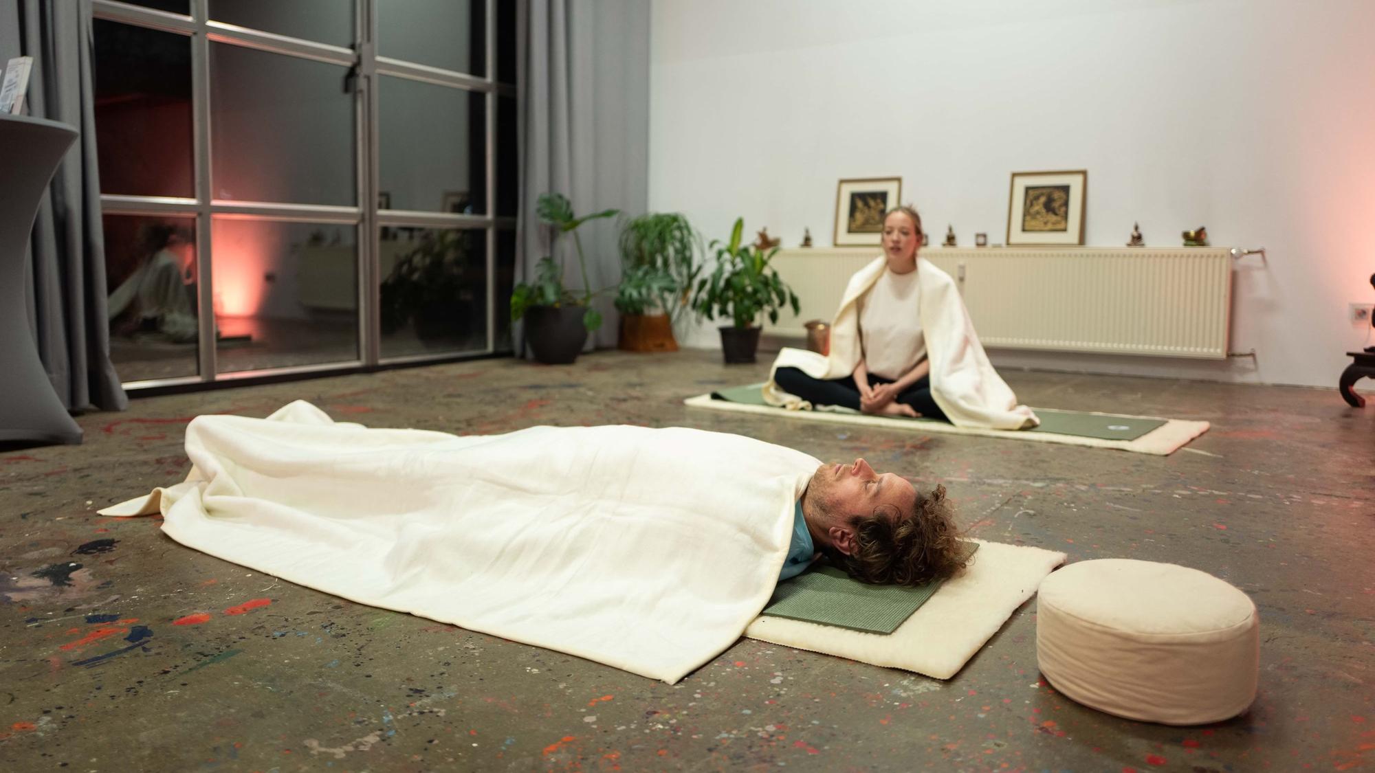 Tom liegt auf dem Rücken auf der Yoga-Matte, über ihm eine weiße Decke. Er hat die Augen geschlossen.
