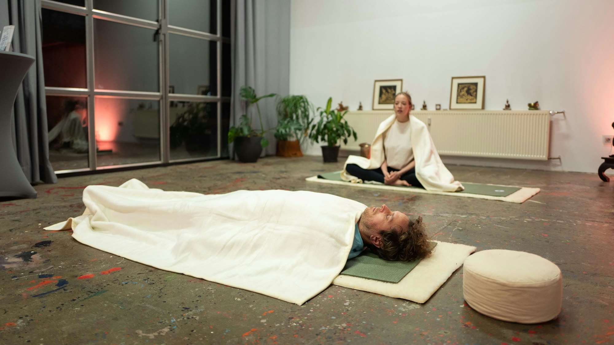 Tom liegt auf dem Rücken auf der Yoga-Matte, über ihm eine weiße Decke. Er hat die Augen geschlossen.