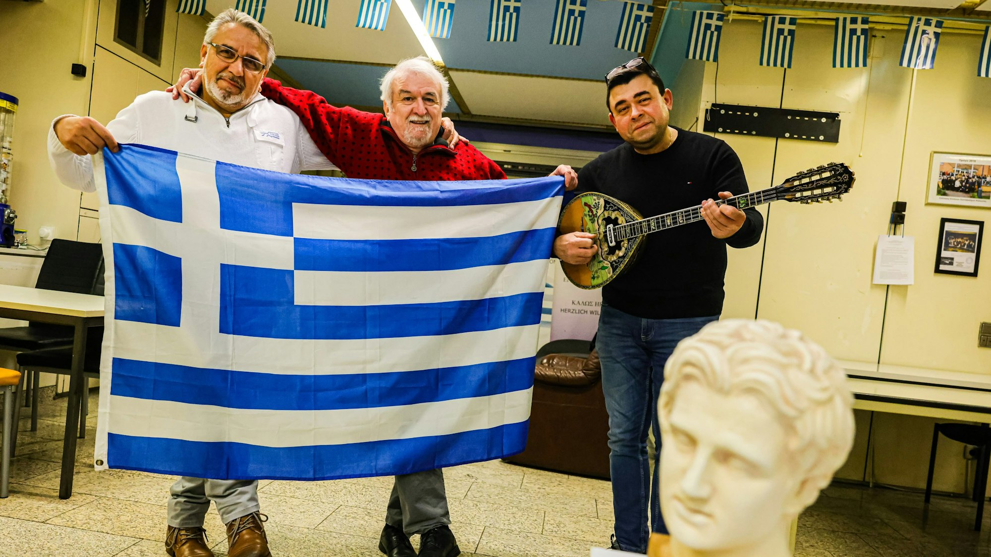 Kostas Giovanoglou (l.) und Vissarion Klissouras (M.) halten eine Griechische Flagge in der Hand, daneben steht Piros Dimitrios mit einem Musikinstrument namens Bouzouki.