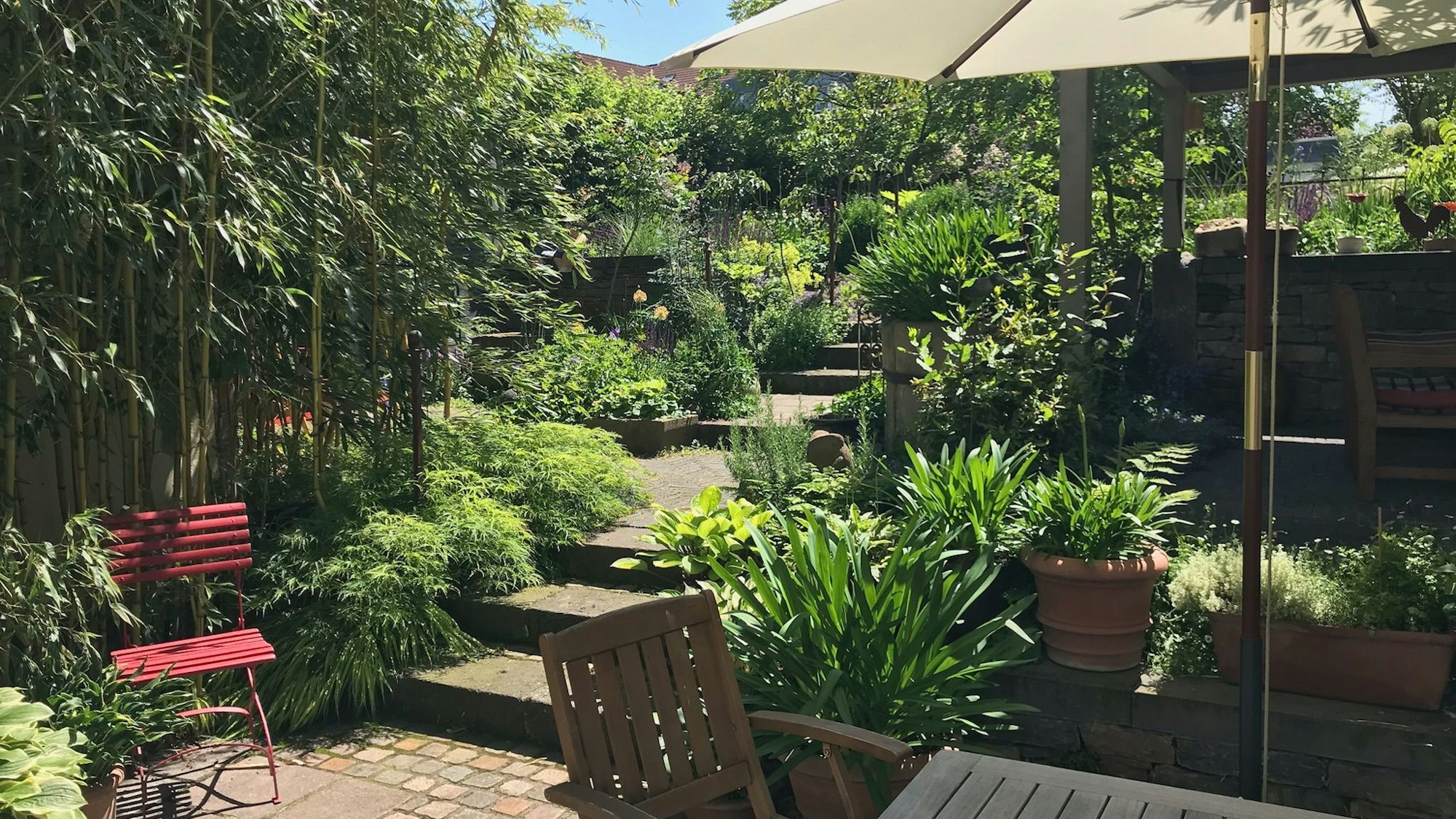 Ein Blick in die grüne Gartenanlage. In der Mitte führt ein Steinweg hoch. Rechts steht ein Holztisch mit Stühlen und einem Sonnenschirm.