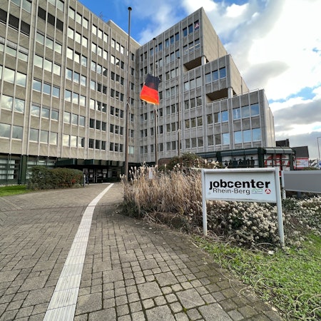 Ein Schild mit der Aufschrift „Jobcenter“ steht vor einem Hochhaus. In dem Hochhaus findet sich das Jobcenter Rhein-Berg.