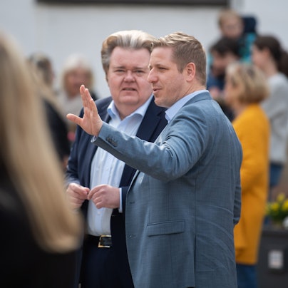 Zwei Männer stehen in einer Gruppe, einer davon gestikuliert mit dem linken Arm.