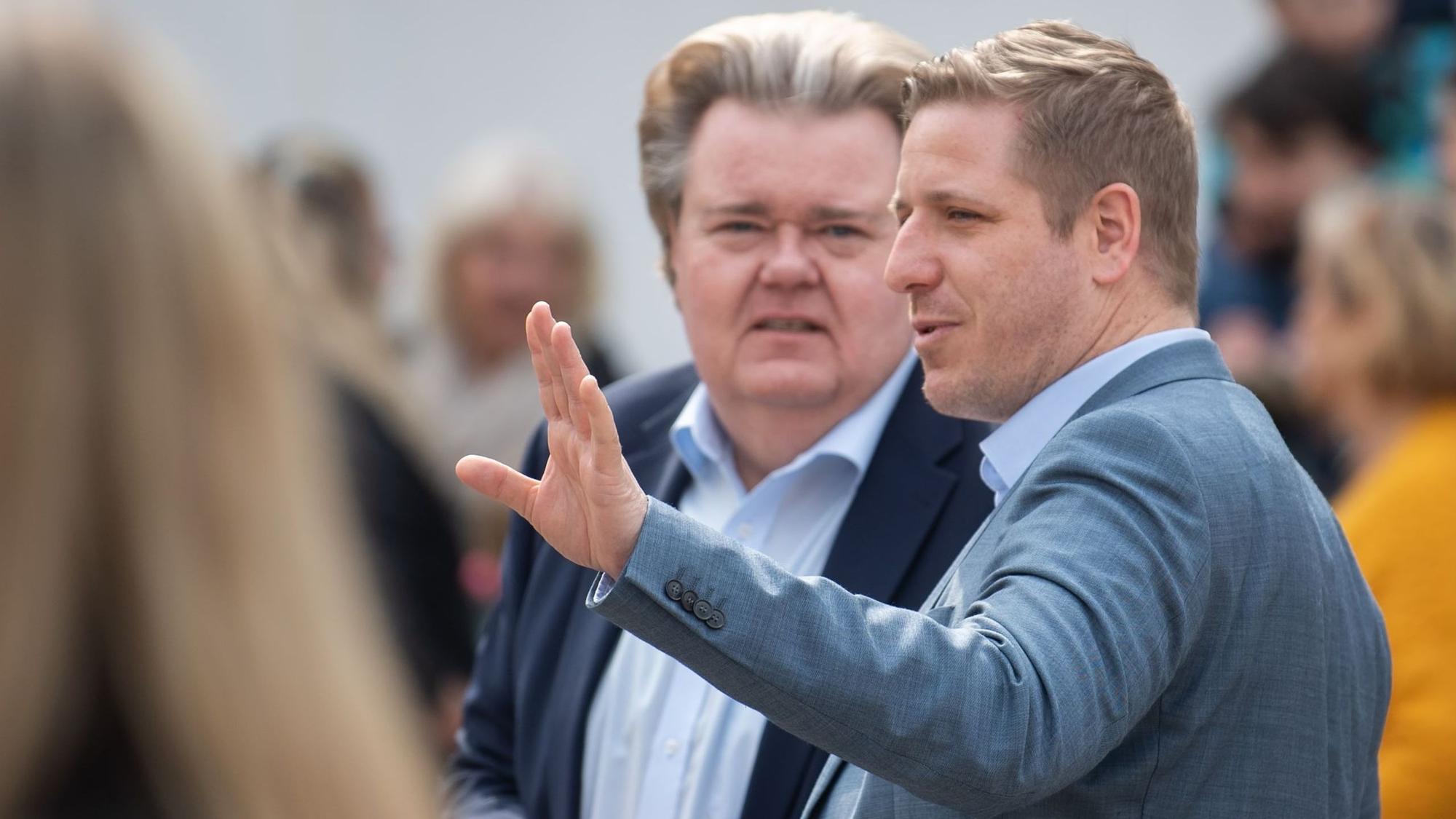 Zwei Männer stehen in einer Gruppe, einer davon gestikuliert mit dem linken Arm.