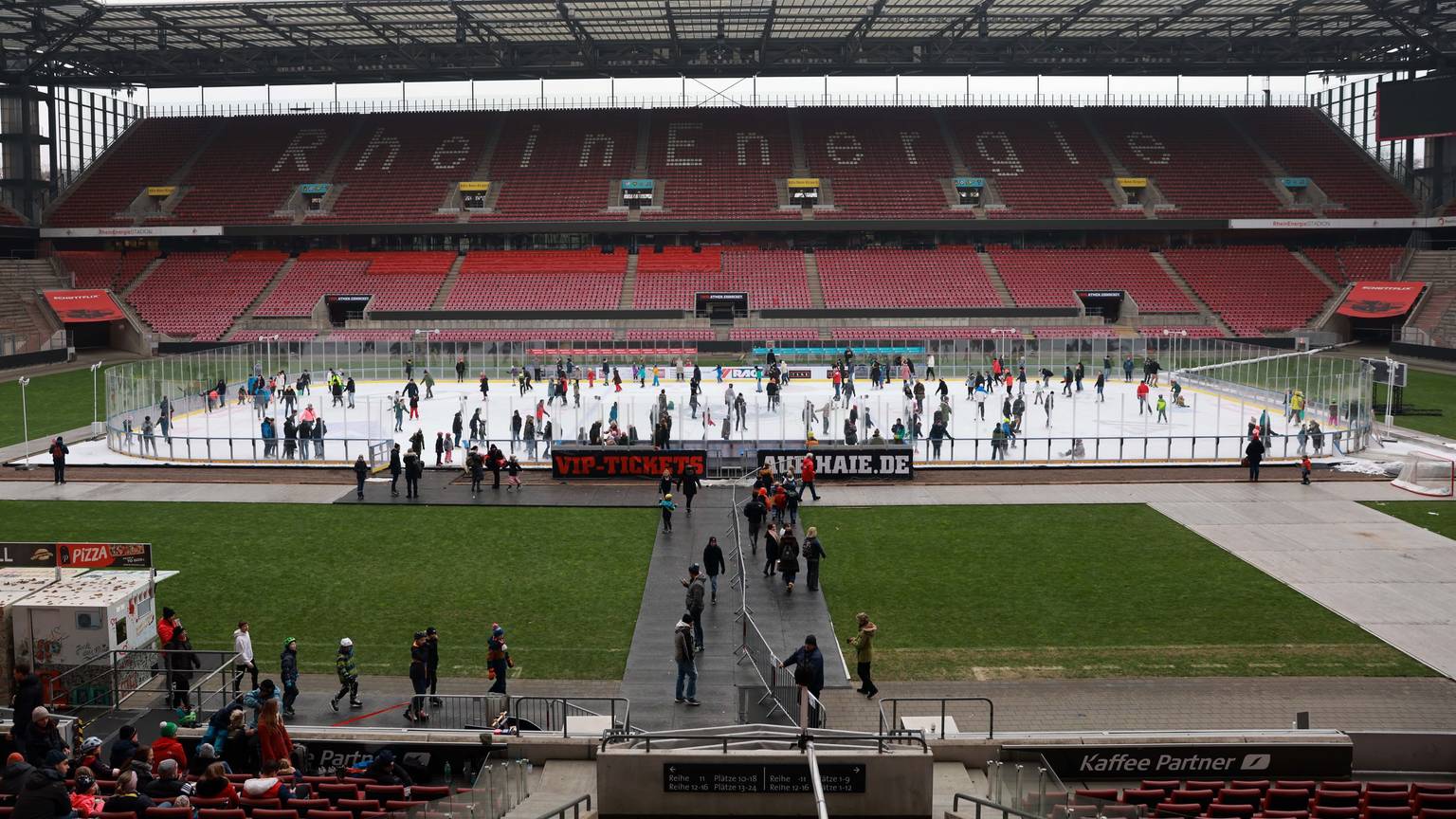 Auf der Eisfläche, die im Rheinenergie-Stadion aufgebaut ist, laufen viele Menschen mit ihren Schlittschuhen. Dahinter ist die leere Tribüne zu sehen.