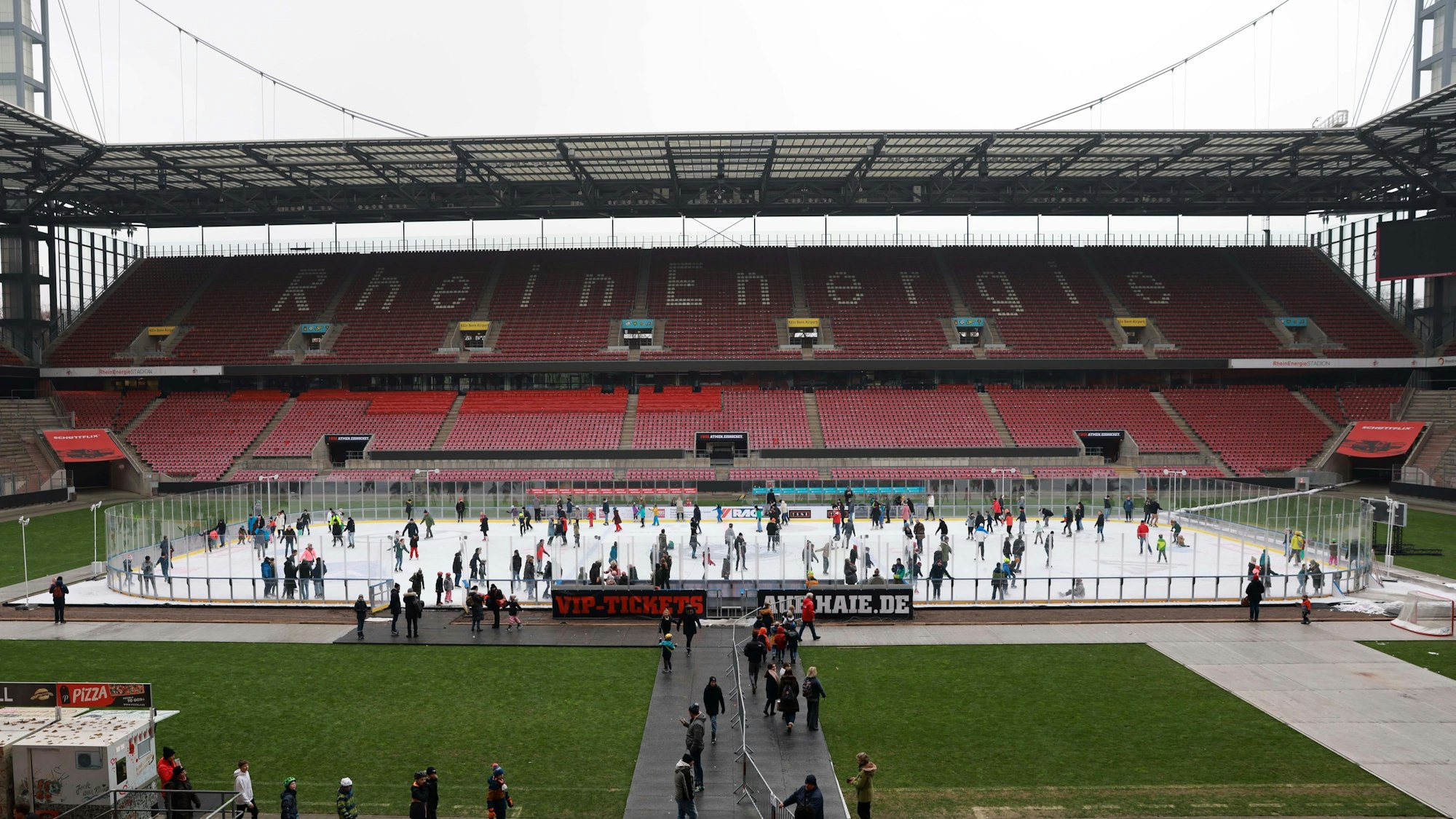 Auf der Eisfläche, die im Rheinenergie-Stadion aufgebaut ist, laufen viele Menschen mit ihren Schlittschuhen. Dahinter ist die leere Tribüne zu sehen.