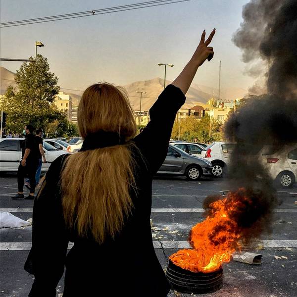 Eine Person mit langen Haaren steht vor einem brennenden Autoreifen und zeigt mit ihrer Hand das Victory-Zeichen.