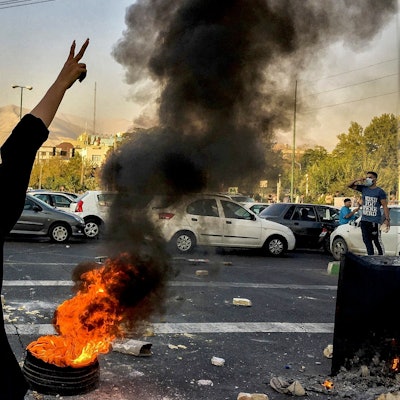 Eine Person mit langen Haaren steht vor einem brennenden Autoreifen und zeigt mit ihrer Hand das Victory-Zeichen.