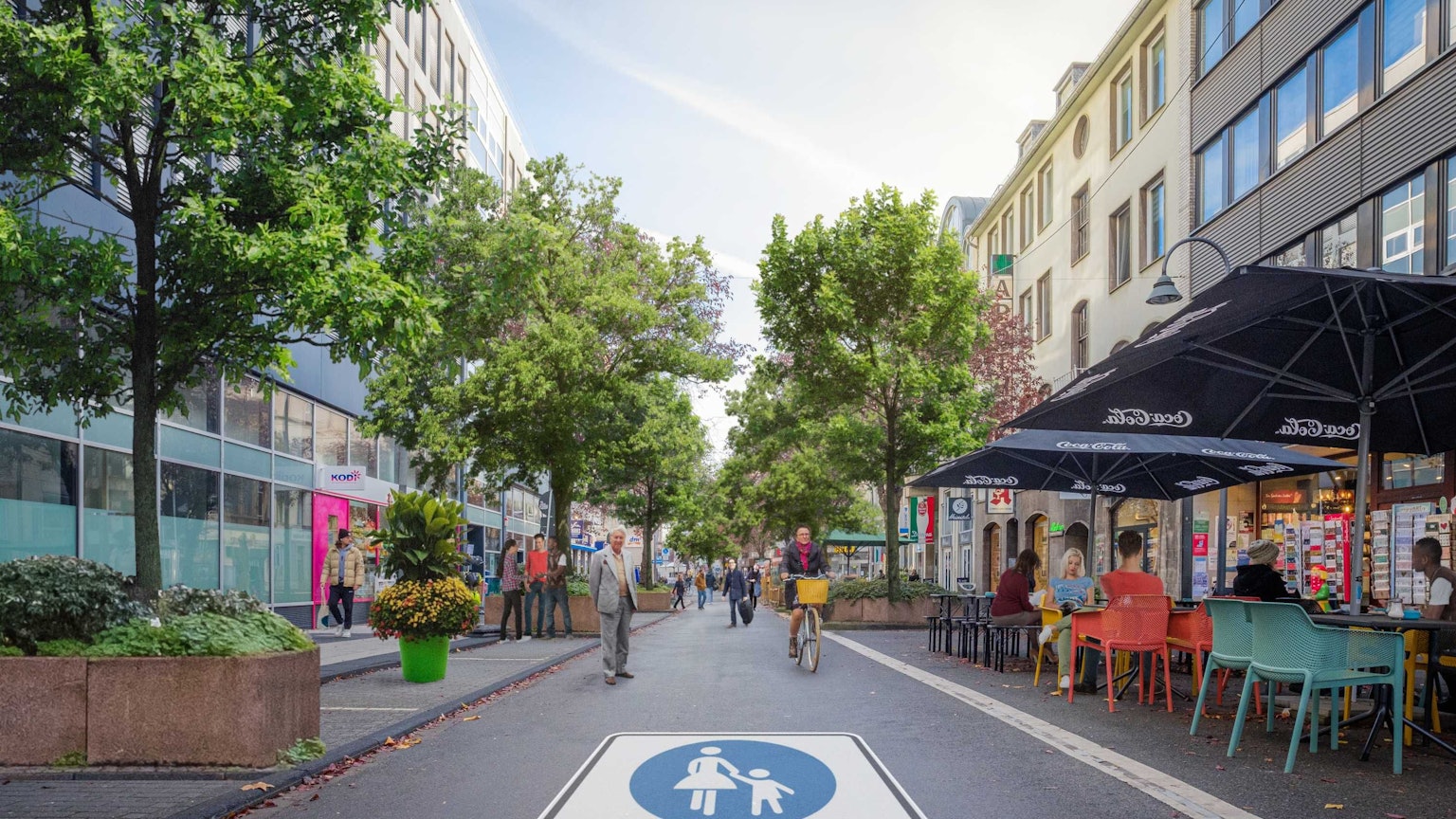 Auf der Straße prangt ein riesiges Piktogramm einer Fußgängerzone. Es sind Radfahrer und Fußgänger unterwegs, in einem Straßencafé sitzen Menschen.