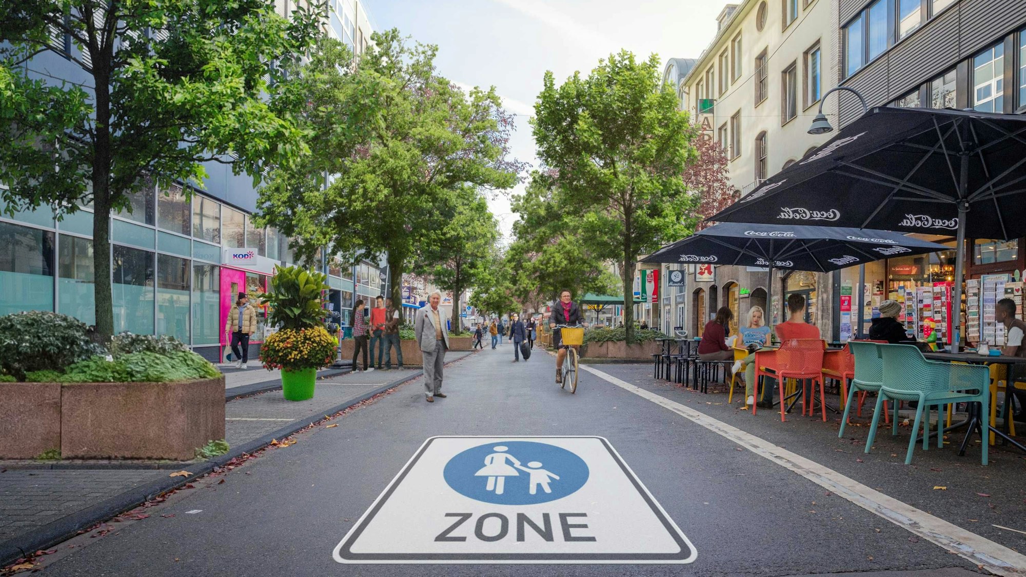 Auf der Straße prangt ein riesiges Piktogramm einer Fußgängerzone. Es sind Radfahrer und Fußgänger unterwegs, in einem Straßencafé sitzen Menschen.