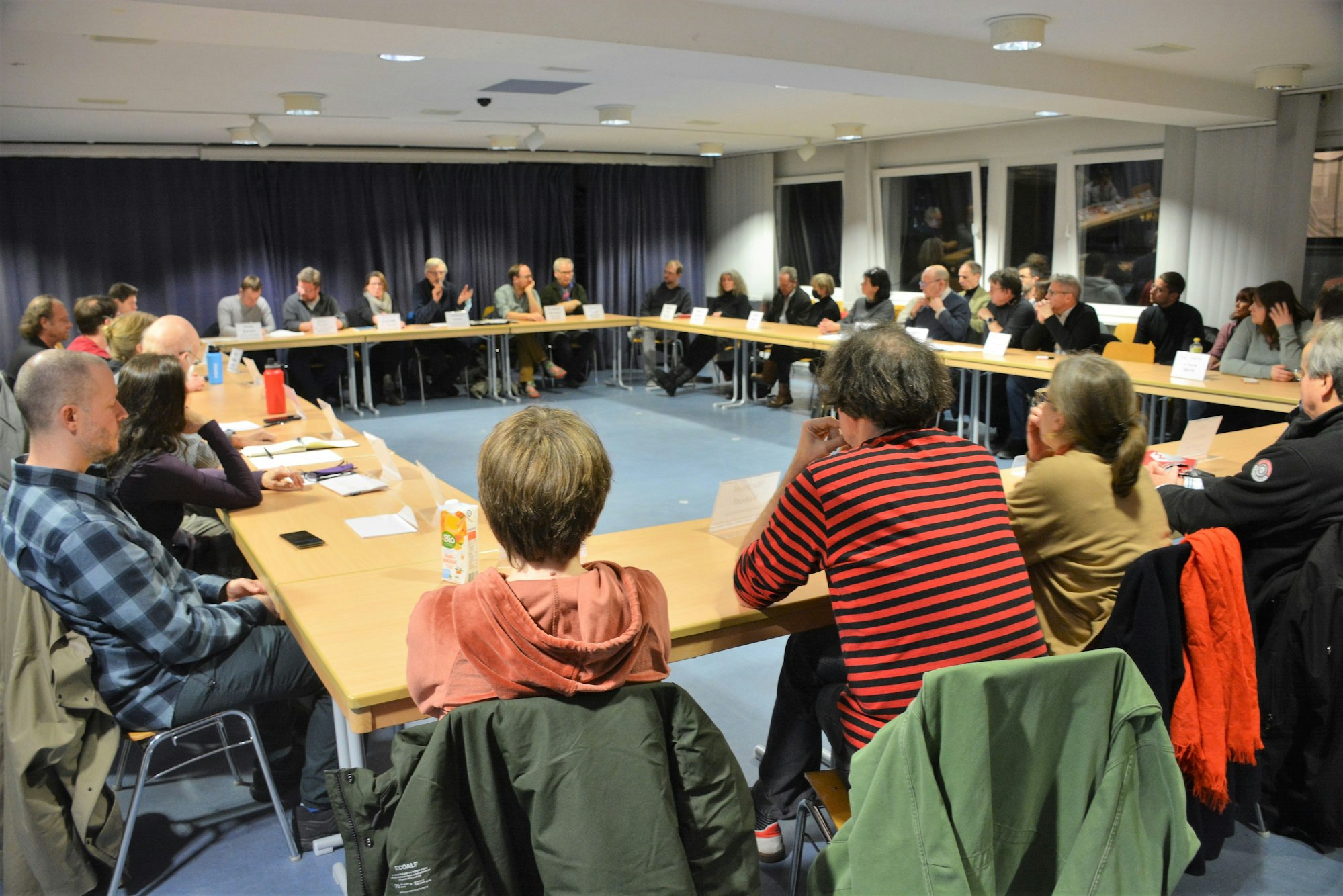 30 Menschen sitzen an in einem großen Rechteck aufgebauten Tischen.
