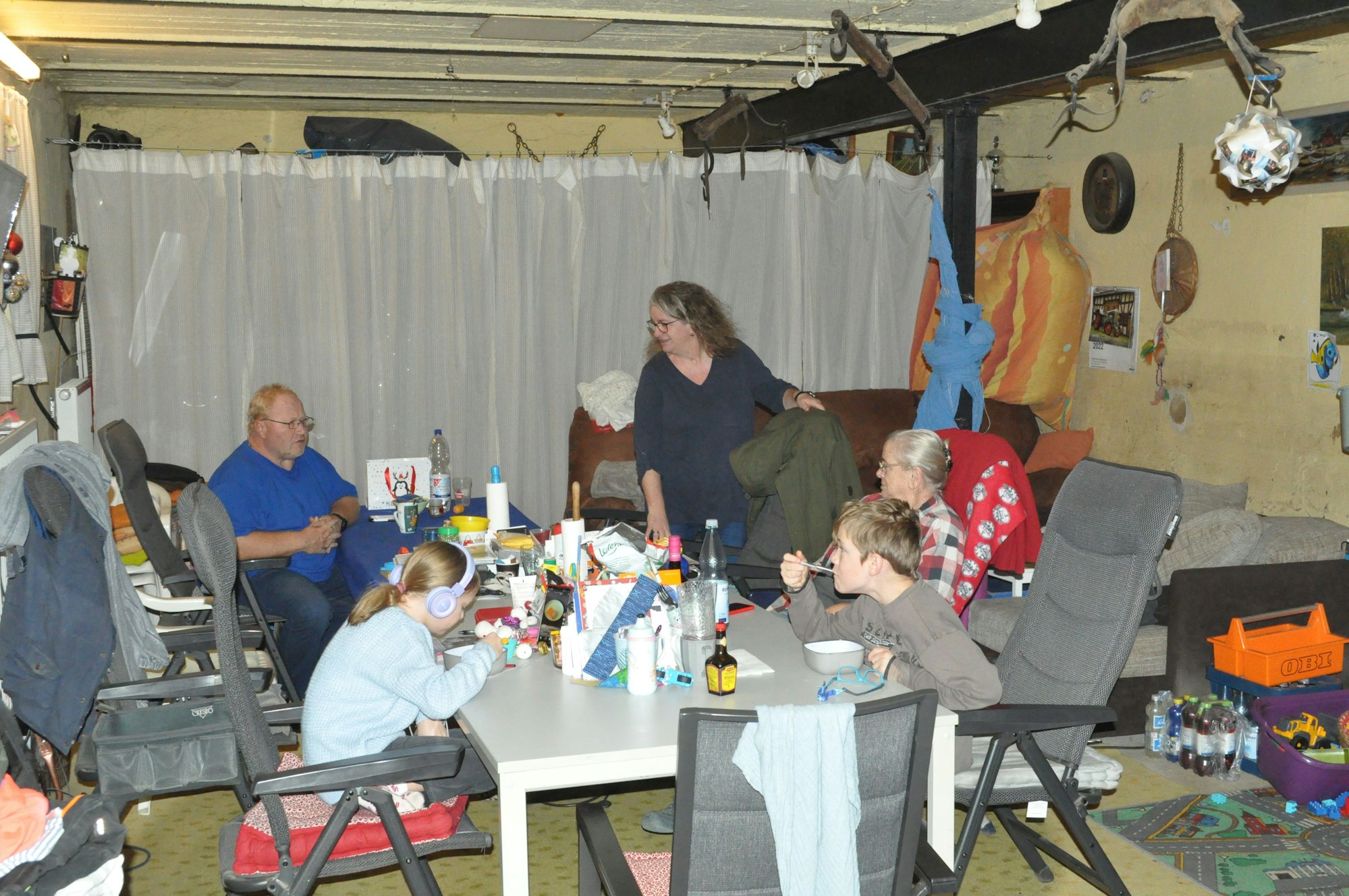 Die Großfamilie Cramer lebt mit neun Personen (im Bild Ludwig und Kirsten mit den Kindern Luis und Lilly sowie Oma Thea) tagsüber im alten Viehstall. Hier wird auch gekocht und gewaschen, was einen Lüfter notwendig macht. Hinter dem Vorhang ruhen die geretteten Überreste von drei Haushalten