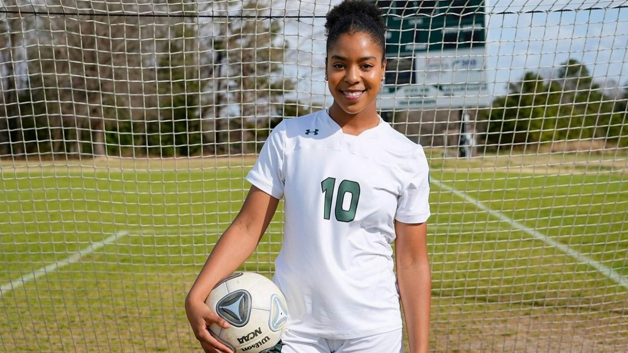 Die Fußballerin Zahra Fepessi trägt die Nummer 10 auf ihrem Trikot und steht in Fußballkleidung und mit einem Ball unter dem Arm vor einem Fußballtor.