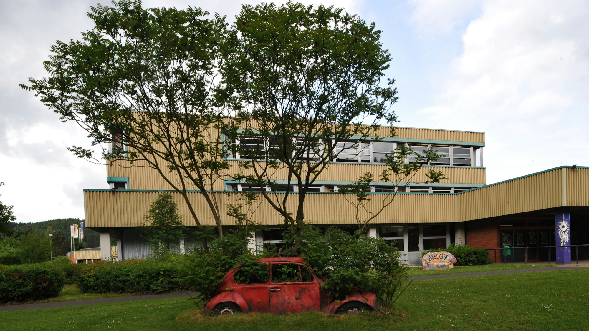 Das Schulzentrum Cyriax in Overath vor Bäumen, davor ein mit Pflanzen bewachsener roter Wagen.