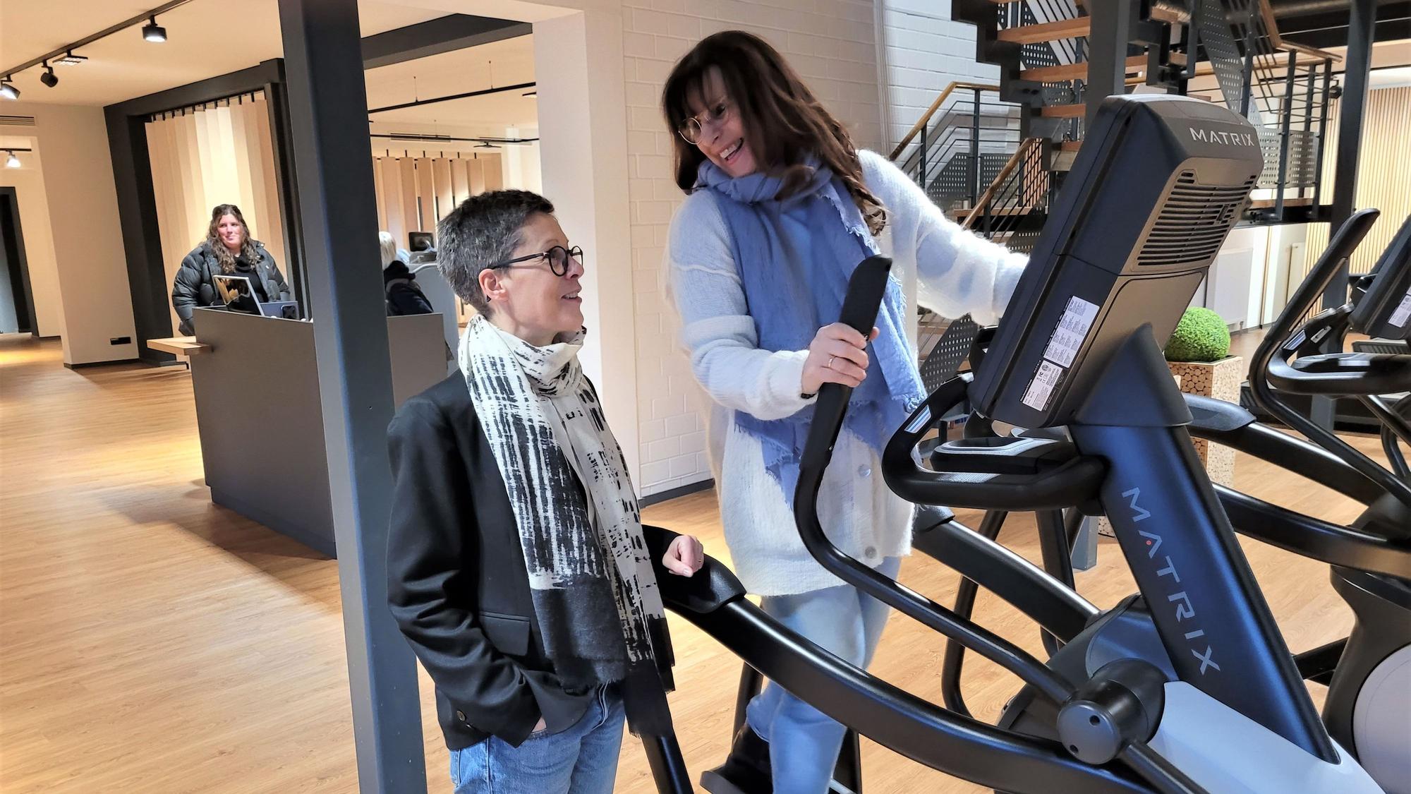 Eine Frau trainiert an einem Fitnessgerät, eine andere schaut ihr zu.