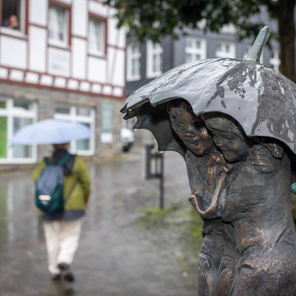 Eine Bronze-Statue aus zwei Personen hat einen Regenschirm über den Köpfen. Im Hintergrund geht eine Person mit Regenschirm.