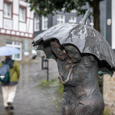 Eine Bronze-Statue aus zwei Personen hat einen Regenschirm über den Köpfen. Im Hintergrund geht eine Person mit Regenschirm.