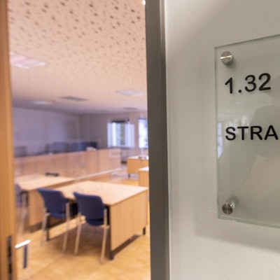 Die Tür eines Gerichtssaals steht einen Spalt offen. Im inneren sind leere Stühle und Tische zu sehen.