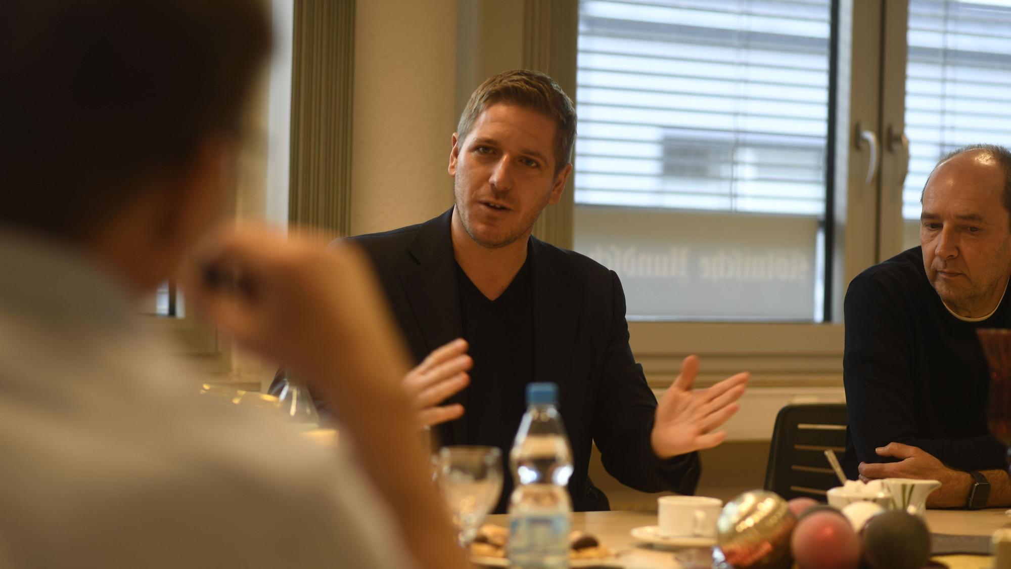 Drei Männer sitzen im Gespräch am Tisch, einer gestikuliert mit den Händen.