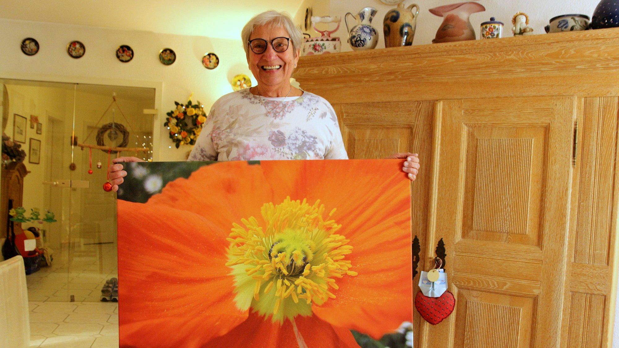Eine mittelalte Frau steht in einem Zimmer. Sie hält ein Bild einer Blume in der Hand.