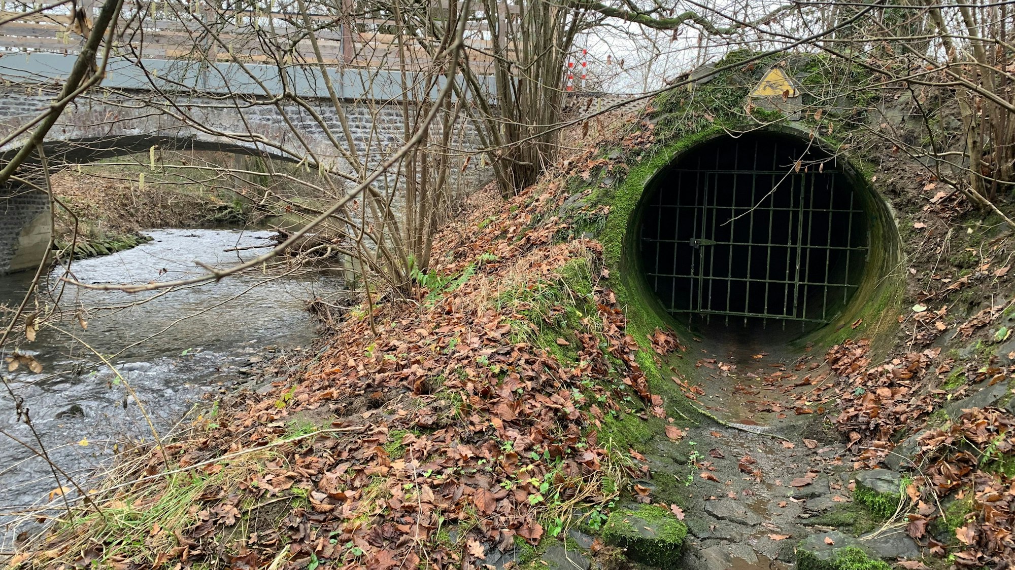 Neben einem Bach, über den eine Brücke führt, ist eine vergitterte Öffnung zu sehen.