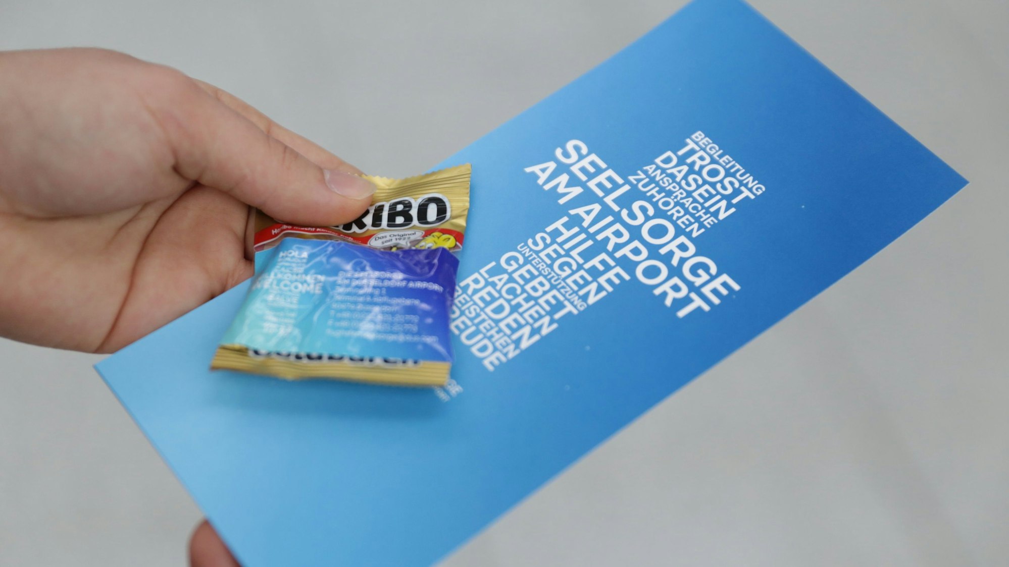 Eine Hand hält eine Tüte Gummibärchen und ein Lesezeichen mit Segensspruch.