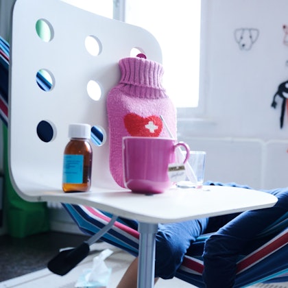 Fieberthermometer, Hustensaft, Wärmflasche und eine Tasse Tee stehen auf einem Stuhl.