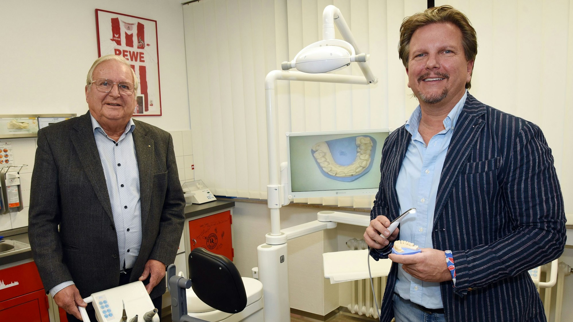 Die Doktoren Wilfried und Sascha Bonness stehen in einem Behandlungszimmer in ihrer Zahnarztpraxis und zeigen moderne medizinische Technik.