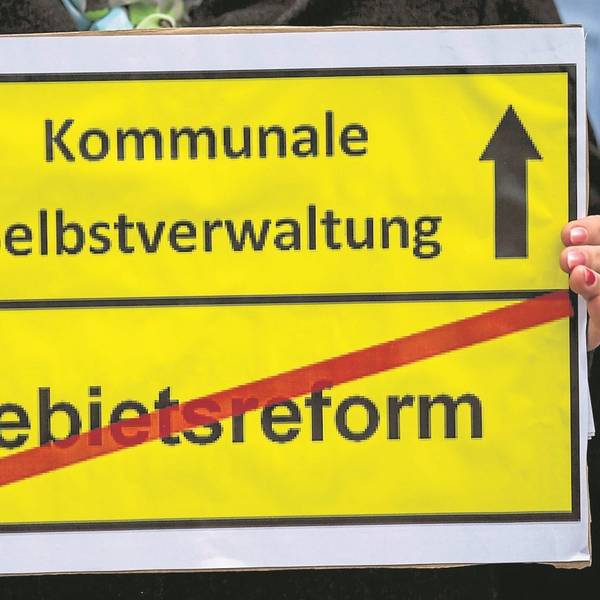 Ein gelbes Schild: Oben die Aufschrift „Kommunale Selbstverwaltung“ mit einem Pfeil nach oben daneben, unten die Aufschrift „Gebietsreform“ mit einem roten Balken durchgestrichen.