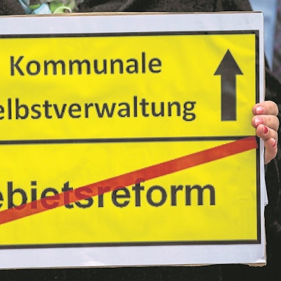 Ein gelbes Schild: Oben die Aufschrift „Kommunale Selbstverwaltung“ mit einem Pfeil nach oben daneben, unten die Aufschrift „Gebietsreform“ mit einem roten Balken durchgestrichen.